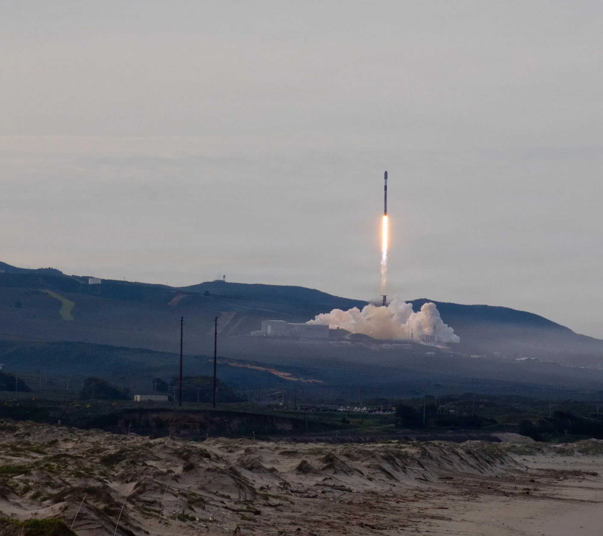 A Falcon 9 rocket climbing from its launch pad at Vandenberg Space Force Base, exhaust clouds spreading below it
