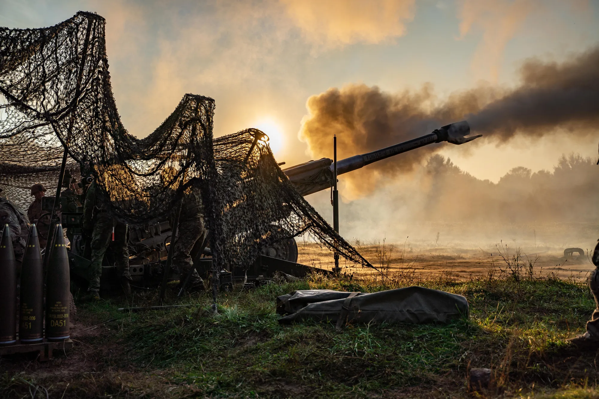 An M777 howitzer firing at dawn from under camouflage netting, muzzle flash and smoke against the rising sun